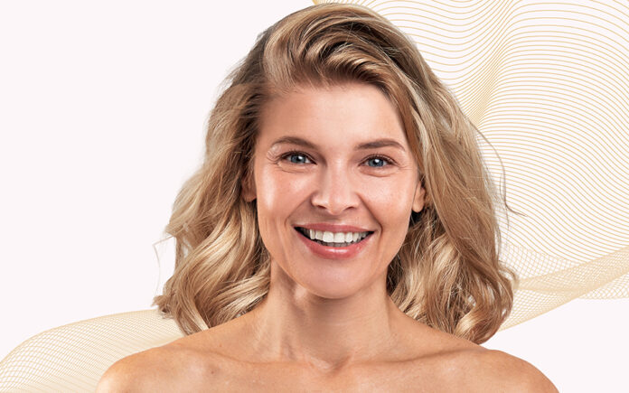 site-industries-cosmetiques Une femme aux cheveux blonds mi-longs sourit à la caméra devant un fond clair orné de lignes ondulées beiges. Elle a le teint clair et est photographiée de la tête aux épaules.