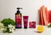site-industries-cosmetiques Um frasco, um tubo e um conjunto de produtos de higiene pessoal da Floral Street estão dispostos sobre uma superfície clara, tendo como pano de fundo flores, meio limão, folhas verdes e talos de ruibarbo.