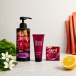 site-industries-cosmetiques Un frasco, un tubo y un estuche de productos de cuidado personal de Floral Street se presentan sobre una superficie clara, con flores, medio limón, hojas verdes y tallos de ruibarbo al fondo.