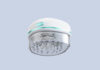 site-industries-cosmetiques Un appareil de massage électronique pour le cuir chevelu de la marque Nuon Medical, de couleur blanche et bleu sarcelle clair, doté d'une base transparente comportant de multiples picots de massage en silicone, présenté sur un fond uni gris clair.