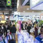 site-industries-cosmetiques Uma multidão numerosa passeia e conversa numa feira profissional ou numa exposição muito animada que decorre num recinto fechado. Os stands e os expositores ladeiam os corredores, e uma bandeira brasileira flutua por cima da multidão. Os visitantes estão vestidos com roupa profissional ou informal.