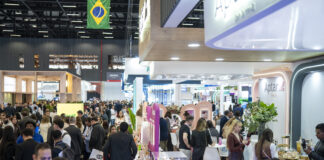2026年FCE展会:参展商数量创纪录,聚焦化妆品行业 site-industries-cosmetiques 在一场热闹非凡的室内专业展会或展览会上,大批人群穿梭往来、交流互动。过道两侧排列着展位和展示架,人群上方飘扬着一面巴西国旗。 参观者们有的身着商务正装,有的则穿着休闲装。.