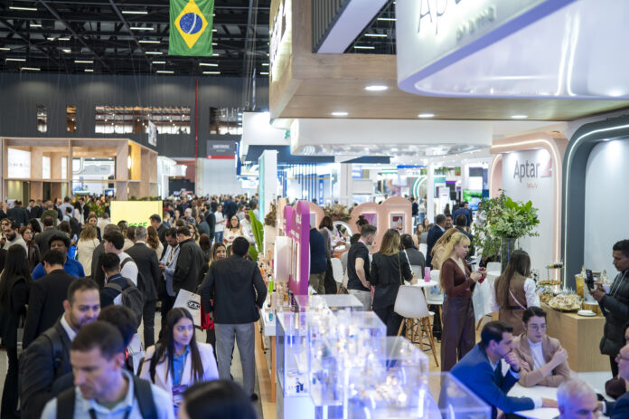 site-industries-cosmetiques Une foule nombreuse se promène et échange dans un salon professionnel ou une exposition très animée qui se tient en intérieur. Des stands et des présentoirs bordent les allées, et un drapeau brésilien flotte au-dessus de la foule. Les visiteurs sont vêtus de tenues professionnelles ou décontractées.