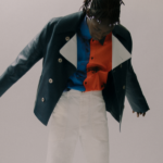 site-industries-cosmetiques Une personne se tient debout à l'intérieur, les bras légèrement écartés. Elle porte une veste vert foncé par-dessus une chemise aux motifs abstraits rouges et bleus, ainsi qu'un pantalon blanc. L'arrière-plan est uni et neutre.