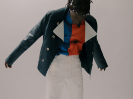 site-industries-cosmetiques Una persona está de pie en el interior, con los brazos ligeramente separados. Lleva una chaqueta verde oscuro sobre una camisa con motivos abstractos rojos y azules, así como unos pantalones blancos. El fondo es liso y neutro.
