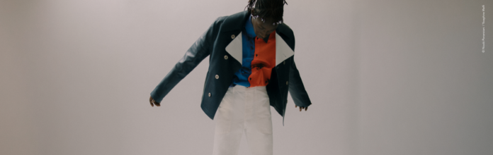 site-cosmetics-industries A person is standing inside, with their arms slightly apart. They are wearing a dark green jacket over a shirt with abstract red and blue patterns, as well as white pants. The background is plain and neutral.