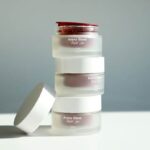 Arena Glass Refill Jar:一款60毫升的玻璃可补充装罐,内含50毫升内胆 site-industries-cosmetiques 一摞三只磨砂玻璃罐,罐身上贴有«Arena Glass Relief 2.0»标签,其中一只白色盖子已被取下,放在一旁,背景为被自然光照亮的素色平面。.
