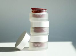 site-industries-cosmetiques 一摞三只磨砂玻璃罐，罐身上贴有«Arena Glass Relief 2.0»标签，其中一只白色盖子已被取下，放在一旁，背景为被自然光照亮的素色平面。.