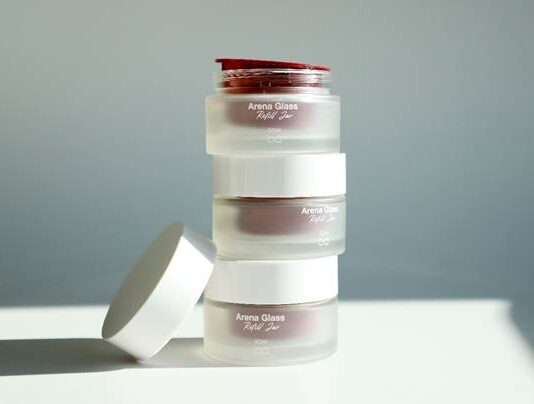 site-industries-cosmetiques Une pile de trois bocaux dépolis portant l'étiquette « Arena Glass Relief 2.0 », dont l'un a son couvercle blanc retiré et posé à côté, sur un fond uni baigné de lumière naturelle.