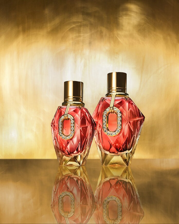 site-industries-cosmetiques Two faceted perfume bottles, red and gold, with gold caps, are arranged side by side on a reflective surface against a gold background. Each bottle is adorned with a decorative oval pendant made of crystal and gold, suspended from its neck.