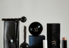 Act Beauty signe des cadeaux‑packagings 2‑en‑1 pour Ella Baché site-industries-cosmetiques Une collection de produits de beauté ou de soins pour hommes, de couleur noire et marron, comprenant un accessoire de massage texturé, des récipients ronds, des cylindres, une brosse et deux étuis en cuir ou en simili-cuir, disposés sur une surface neutre avec un fond uni.