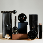 Act Beauty signe des cadeaux‑packagings 2‑en‑1 pour Ella Baché site-industries-cosmetiques Une collection de produits de beauté ou de soins pour hommes, de couleur noire et marron, comprenant un accessoire de massage texturé, des récipients ronds, des cylindres, une brosse et deux étuis en cuir ou en simili-cuir, disposés sur une surface neutre avec un fond uni.