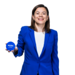 Beiersdorf Appoints Isabelle Schang to Lead Marketing for France and Benelux site-cosmetics-industries A woman wearing a blue blazer and a white shirt is standing in front of a white background, smiling, holding a blue jar in her left hand that reads «NIVEA Crème,» with the logo clearly visible.