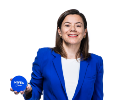 site-cosmetics-industries A woman wearing a blue blazer and a white shirt is standing in front of a white background, smiling, holding a blue jar in her left hand that reads «NIVEA Crème,» with the logo clearly visible.