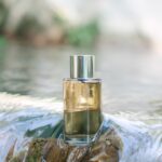 Coverpla étend la rechargeabilité, du flacon aux recharges site-industries-cosmetiques Un flacon de parfum en verre transparent, muni d'un bouchon argenté, est posé sur un rocher partiellement immergé dans un cours d'eau peu profond. L'arrière-plan est légèrement flou, baigné par la lumière naturelle extérieure.