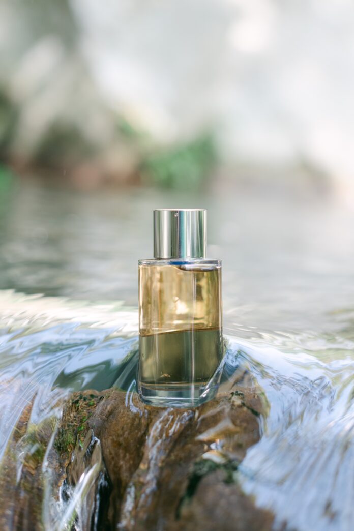 site-industries-cosmetiques Un flacon de parfum en verre transparent, muni d'un bouchon argenté, est posé sur un rocher partiellement immergé dans un cours d'eau peu profond. L'arrière-plan est légèrement flou, baigné par la lumière naturelle extérieure.