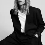 Rabanne confía la dirección creativa a Olivier Rousteing site-industries-cosmetiques Una persona con el pelo largo y ondulado está sentada en un taburete; lleva una chaqueta de traje negra, una camisa blanca y unos pantalones negros. Tiene una mano en el bolsillo y lleva un reloj. El fondo es liso y claro.