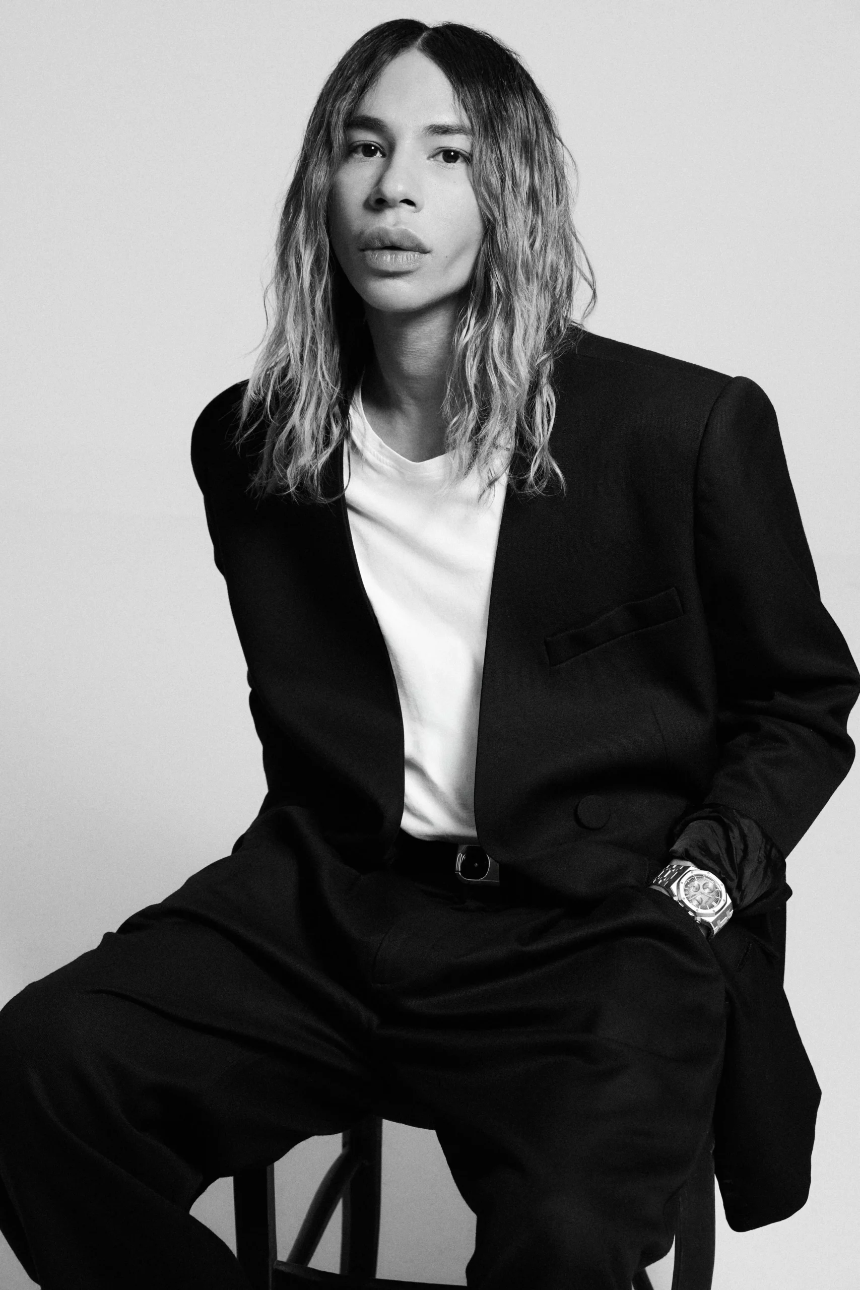 site-industries-cosmetiques Une personne aux cheveux longs et ondulés est assise sur un tabouret ; elle porte une veste de costume noire, une chemise blanche et un pantalon noir. Elle a une main dans la poche et porte une montre. L'arrière-plan est uni et clair.