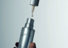 site-industries-cosmetiques 一只手正握着一瓶Dermalogica品牌的«Futurecode Booster»银色瓶装产品。瓶盖已被取下，瓶口处的滴管正滴出一滴产品。背景为素色。.