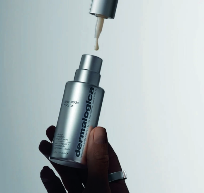 site-industries-cosmetiques Uma mão segura um frasco prateado de «Futurecode Booster» da Dermalogica. A tampa está retirada e um conta-gotas situado na parte superior do frasco dispensa uma gota do produto. O fundo é neutro.